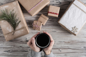 Female with cup of coffee packing gifts. Cardboard box in craft paper, christmas rope and tree on the rustic wood planks background. DIY.