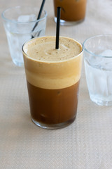 Cold frappe coffee with ice cubes on glass with black straw served on the table in caffeteria. Ice coffee in a tall glass with a lot of foam as cold summer drink.