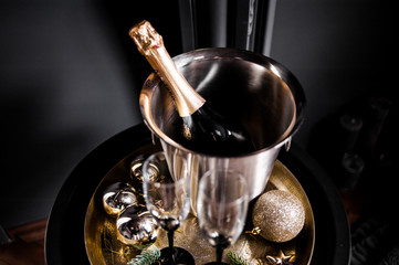 Champagne bottle in freezer bucket with glasses on dark background.