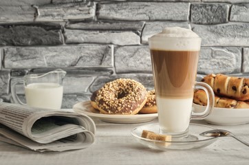 Conceptual delicious sweet breakfast. Latte macchiato coffee  with sweet donuts with a hole and morning newspaper.