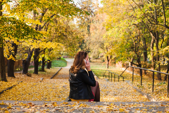 The beautiful young girl sitting on the stairs in the autumn park. Brunette in a leather jacket with a phone in her hands.