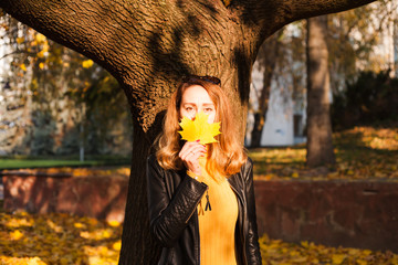 Beautiful young woman in a leather jacket covering her face with a yellow autumn leaf in the autumn...