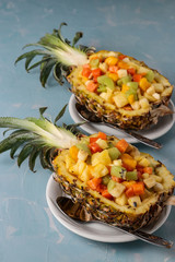 Tropical fruit salad in pineapple halves on white plates on a light blue background, vertical orientation