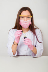  A girl in gloves and a white coat sits at a table with varnishes and holds a bottle of varnish and a brush for nails in her hands. Studio photo on a white background.