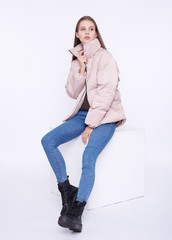 Young girl in a jasmine down jacket sitting on a white cube isolated on a white background. © Alexey