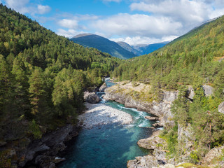 Naklejka na ściany i meble View on azure Rauma river canyon at Romsdalen valley with snow capped peaks of mountains, rocks and green forest. Blue sky white clouds background. Norway summer landscape scenery