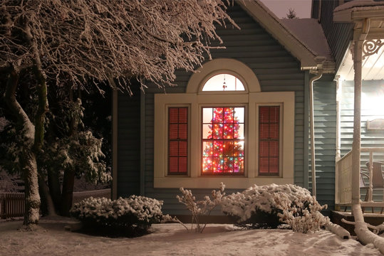 Christmas and New Year background. Night scene with decorated Christmas tree glowing in a room of the private house, view from outdoor with fresh snow on a front yard in foreground.