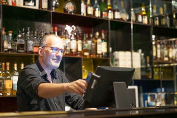 Professional elderly bartender in eyeglases registration of a new order by a cash register. Service concept. 