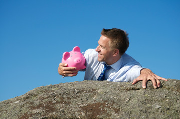 Smiling businessman holding onto a trusty pink piggy bank as he reaches the top of a rocky cliff