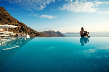 Solitary man sitting on the edge of an infinity pool looking out over the scenic Mediterranean view of the Santorini caldera, Greece