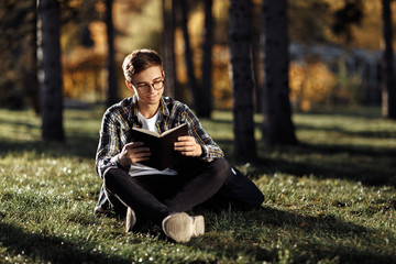 Naklejka na ściany i meble Smile young male student in casual outfit reading a book while sitting on the grass at the park.