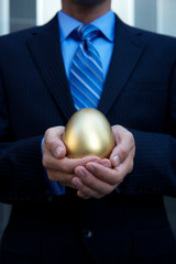 Unrecognizable businessman holding a golden egg cradled in his hands standing outdoors in a dark suit in front of a modern business background