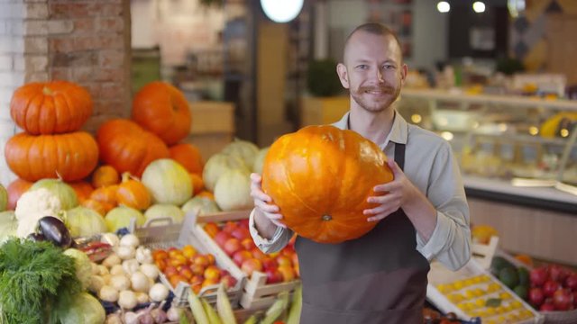 Waist up shot of cheerful mid-aged Caucasian farmer in apron holding ripe pumpkin and smiling at camera while standing beside shelves with fruit and vegetables in food market