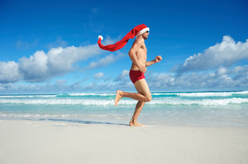 Thin Santa with extra long hat running in red swimming briefs on the shore of a beach in a tropical Christmas fitness regime