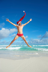 Thin Santa with extra long hat jumping in red swimming briefs on the shore of a beach in a tropical Christmas celebration