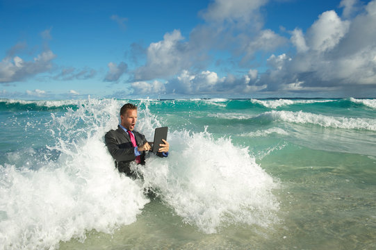 Overwhelmed young businessman in a full suit having a rough time using his tablet computer to surf the web in crashing waves on the shore of a tropical beach