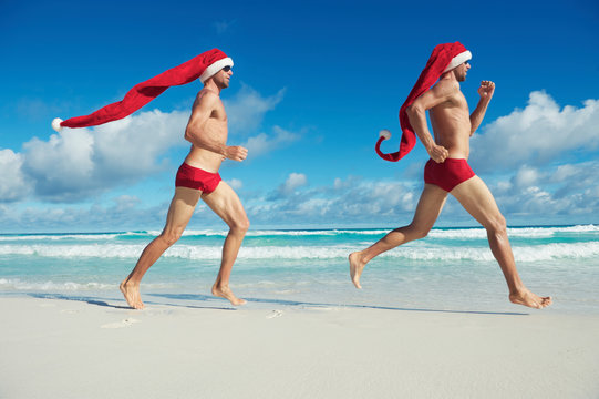 Twin Santas with extra long hats running in red swimming briefs on the shore of a beach in a tropical Christmas fitness regime