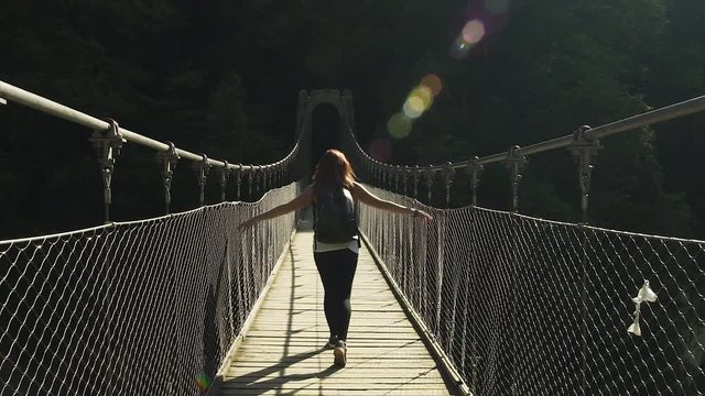 Female Explorer redhead girl walking, hiking, crossing beautiful bridge of Holzarte Footbridge, wooden metal grey bridge, black backpack. 4K UHD. Hands moving along grids, beautiful sun flare.