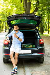 Smiling man with paper cup of coffee sitting in car trunk and looking at camera