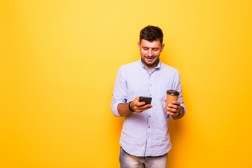 Portrait of beautiful man holding cell phone and takeaway coffee isolated over yellow background