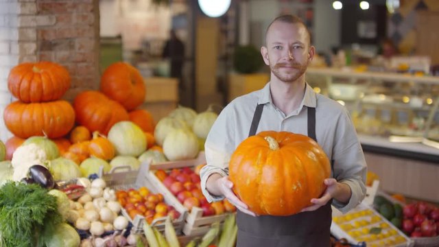 Waist up shot of mid-aged Caucasian salesman in apron holding large pumpkin, looking at camera and smiling while standing beside shelves with fruit and vegetables in food market