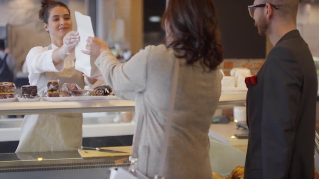 Young beautiful saleswoman in apron and disposable gloves giving paper bag with products to customers and saying bye to them while working at deli department in food mall
