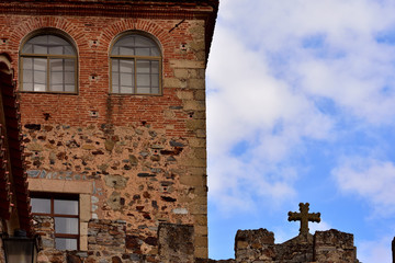 Historic building of the Spanish city of Cáceres next to a stone cross cut into the blue sky.