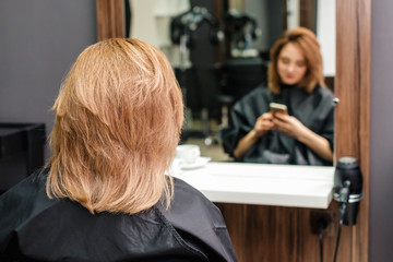 Young woman with new hairstyle is sitting in front of the mirror in the hair salon back view.