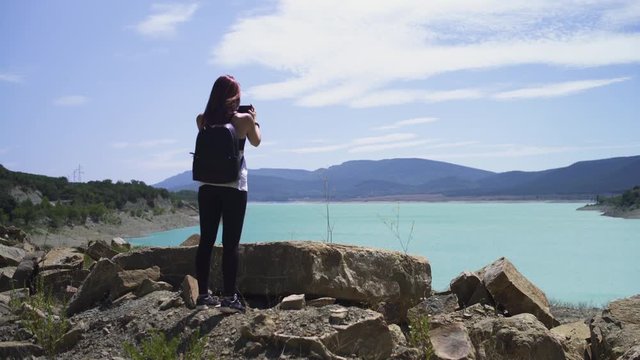 Happy lonely explorer redhead girl standing, hiking at beautiful blue turquoise lake of Yesa, black backpack, during sunny summer in Spain. 4K UHD. Taking picture with smartphone.