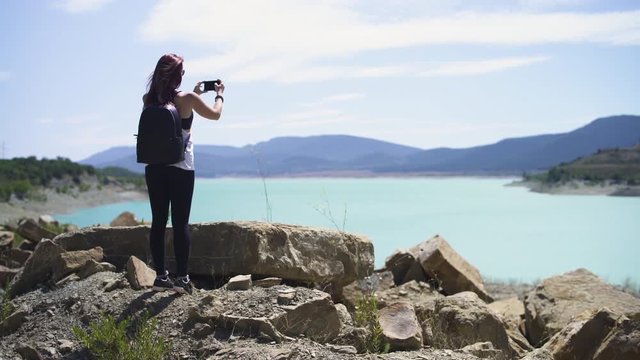 Happy smiling explorer redhead girl standing, hiking at beautiful blue turquoise lake of Yesa, black backpack, during sunny summer in Spain. 4K UHD. Taking picture with smartphone.