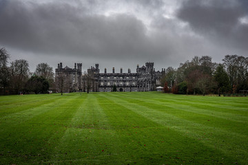Fototapeta na wymiar KILKENNY, IRELAND, DECEMBER 23, 2018: Kilkenny Castle seen from the huge garden on a dramatic cloudy day with dry leaves over the green grass while people run beside it.