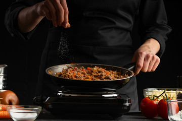 Chef fries vegetables with meat for cooking a delicious dish. Salt with salt. On a black background.