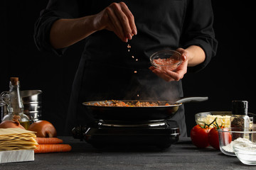 Chef fries vegetables with meat for cooking a delicious dish. Salt with salt. On a black background.