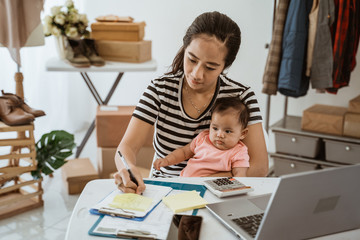 Fototapeta na wymiar Smiling working mother with her baby taking note of orders from customers. Dropshipping business owner working in her office.