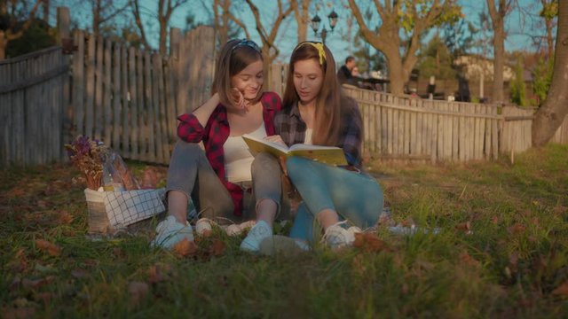 Couple of romantic girls reading books on picnic time at sunset. Cute attractive woman reading a story for her girlfriend enjoying relaxation leisure activity together.