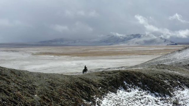 Aerial, drone shot, panning around snowy mountains, revealing a man horseback riding, on hills in the Altai mountains, on a cloudy day, near Bayan-Olgii, in Mongolia