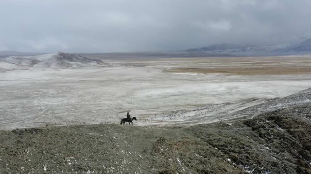 Aerial, drone shot, panning towards a nomad man horseback riding, on snowy hills in the Altai mountains, on a cloudy day, near Bayan-Olgii, in Mongolia