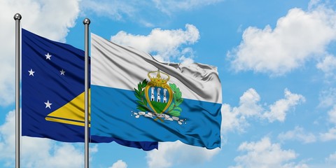 Tokelau and San Marino flag waving in the wind against white cloudy blue sky together. Diplomacy...