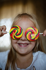 Funny child with candy lollipop, lifestyle photo of happy blonde little girl eating colorful sugar lollipop at home