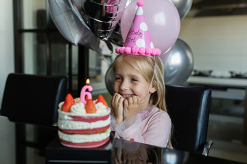 Indoor shot of pretty joyful little girl with blonde hair blowing out the candle, celebrate 6 years old birthday, wear fashionable dress, have excited expressions. Happy childhood concept