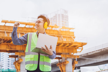 Image outside the industrial construction engineers in yellow protective ear muff discuss new project while using laptop and happy smile on the open building site near the crane.