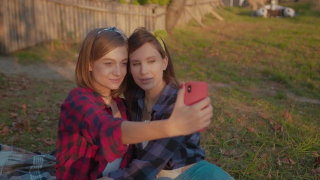 Charming young affectionate women taking nice selfies on smartphone looking on camera staying on pleasant fall picnic in the park. Girls on date.