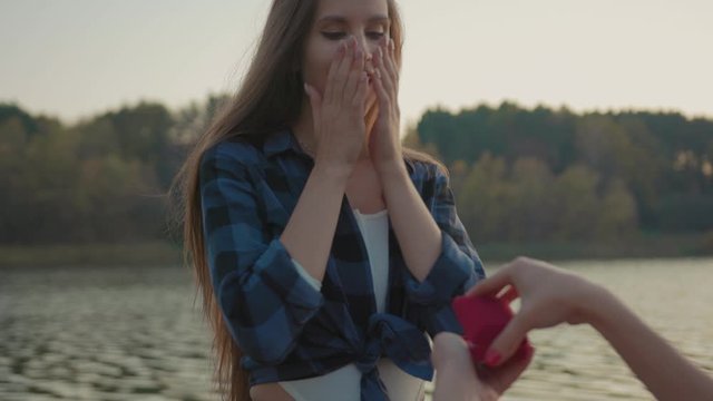 Charming young woman receiving exciting marriage proposal with ring outdoors at lake. Amazed precious girl accepting her girlfriends hand in marriage on romantic date in nature.