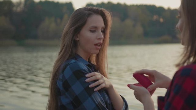 Woman proposing to her beloved sweetheart with engagement ring at beautiful lake. Excited adorable girl accepting her girlfriend marriage proposal hugging in nature outdoors.
