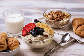 full Breakfast of oatmeal with berries and fruit, croissants, glass of milk, muesli with nuts on light gray background