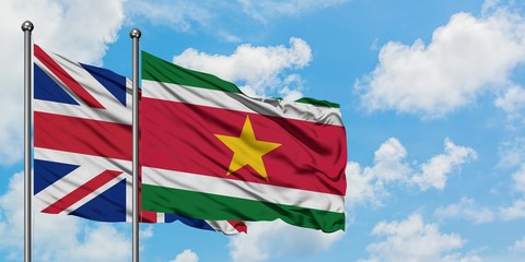 United Kingdom and Suriname flag waving in the wind against white cloudy blue sky together....