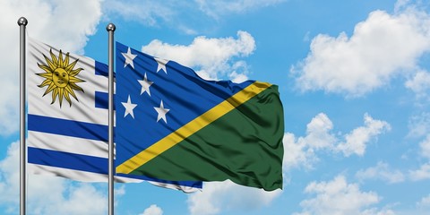 Uruguay and Solomon Islands flag waving in the wind against white cloudy blue sky together....