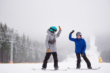 Smiling woman and man giving high five to each other. Happy couple standing on snowboards on snow-covered slope with chairlift on side along coniferous forest. Dense foggy mountain views on background
