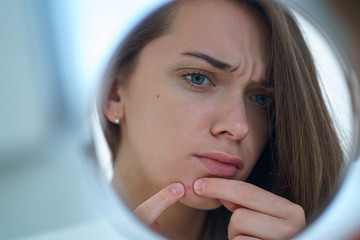 Upset stressed sad acne woman with problem skin squeezes pimple at home using a small round mirror