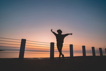 Fototapeta premium Silhouette of one lonely free carefree traveler woman in hat with open outstretched arms standing on sea pier at sunset. Happy freedom concept and enjoying life